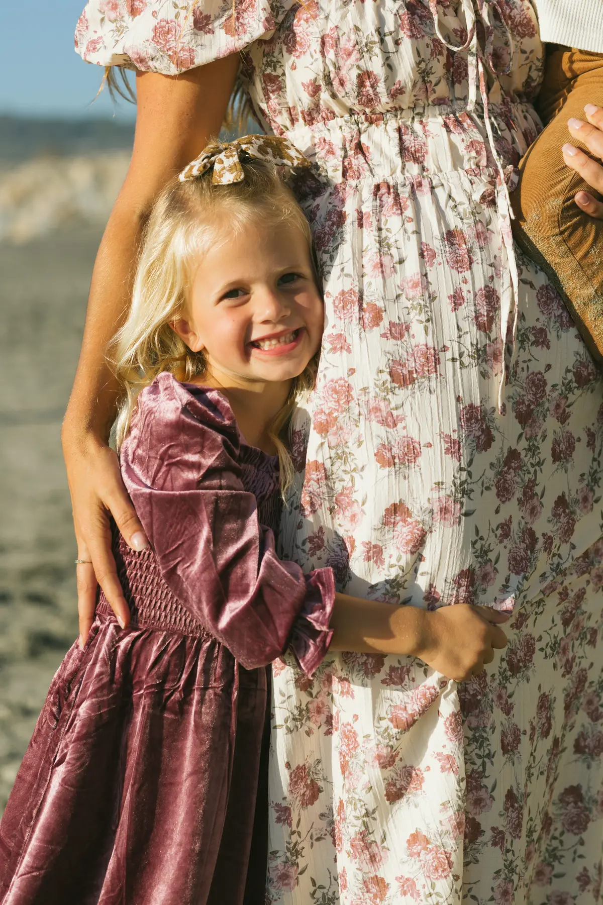 Little girl in a velvet holiday dress hugging her mom’s floral skirt