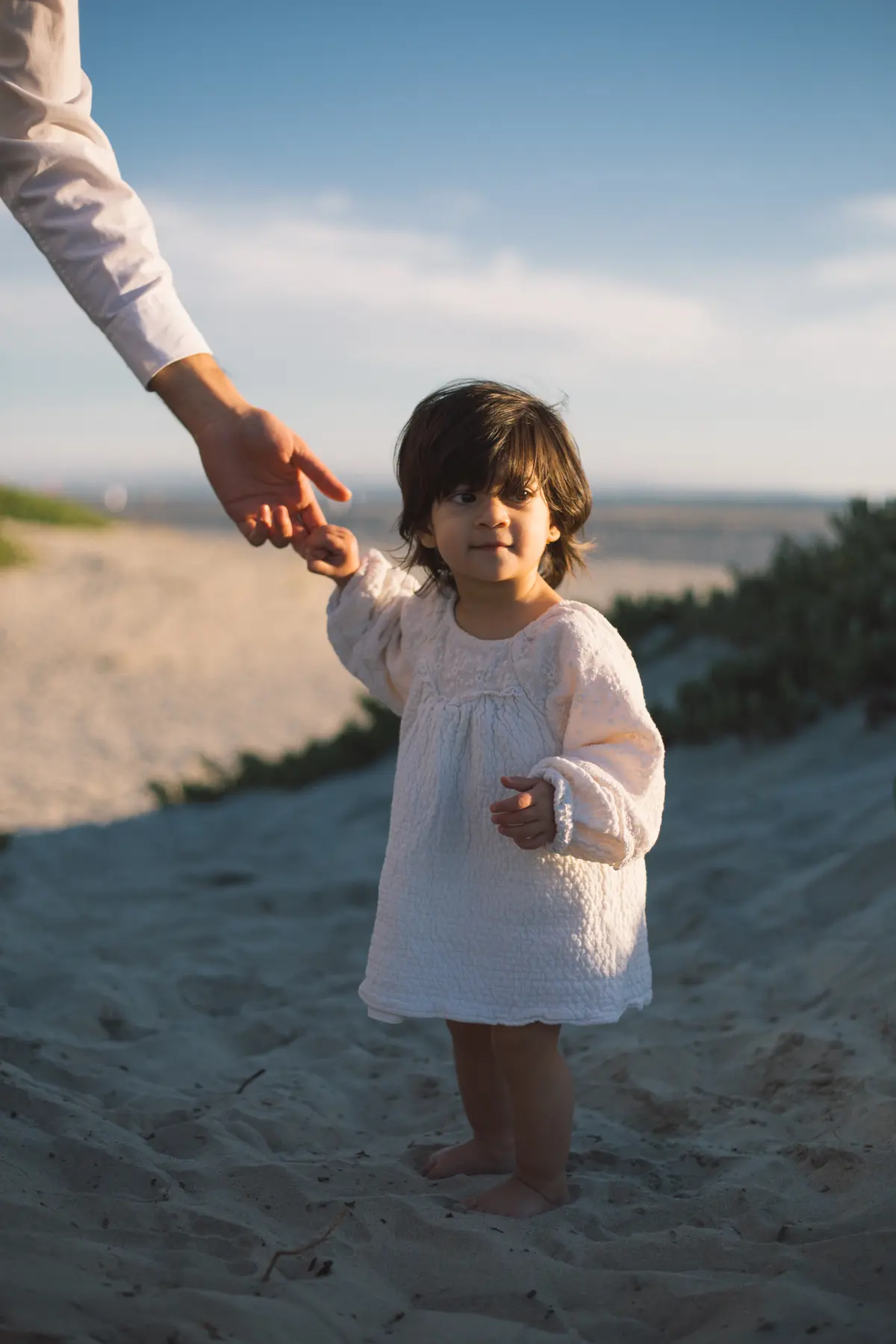 Toddler in a white dress reaching for her parent’s hand on the beach at sunset